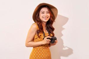 Portrait of cheerful smiling young woman taking photo with inspiration and wearing summer dress. Girl holding retro camera. Model posing on beige background in hat