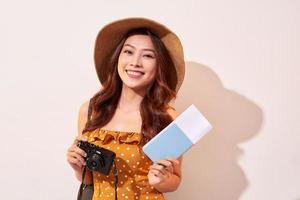 retrato de una joven feliz con sombrero sosteniendo una cámara y mostrando el pasaporte mientras se encuentra aislada sobre un fondo beige foto