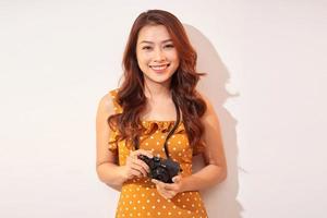 Portrait of cheerful smiling young woman taking photo with inspiration and wearing summer dress. Girl holding retro camera. Model posing on beige background in hat