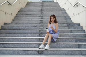 Cheerful young Asian woman holding takeaway coffee cup, using mobile phone photo