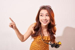 Tourist standing isolated on beige background and joyfully pointing to the sky. Lady using camera while travel. Independent travel in Asia concept. photo