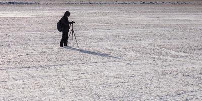 photographer with a tripod in snowy field takes pictures of winter landscape, footprints in snow