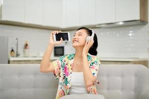 Beautiful emotional pleased young woman on sofa at home listening music with headphones holding mobile phone singing. photo