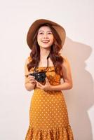 Portrait of cheerful smiling young woman taking photo with inspiration and wearing summer dress. Girl holding retro camera. Model posing on beige background in hat