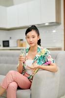 Happy woman taking vitamin pill holding glass sitting on a couch at home photo