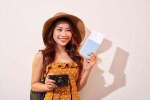 retrato de una joven feliz con sombrero sosteniendo una cámara y mostrando el pasaporte mientras se encuentra aislada sobre un fondo beige foto