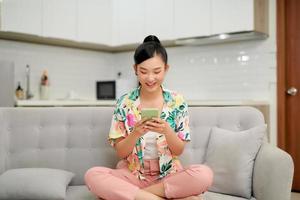 Smiling teenager girl relax sit on sofa at home using cellphone texting chatting with friend photo