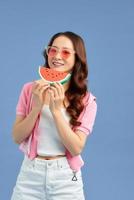 Cheerful young Asian woman holding watermelon and wearing sunglasses over blue background. photo