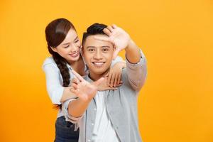 Happy young asian couple looking through a finger frame and smiling while standing isolated on yellow photo
