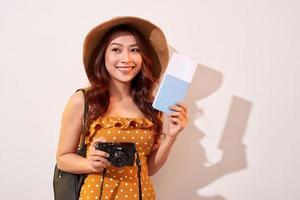 Portrait of a happy young woman in hat holding camera and showing passport while standing isolated over beige background photo