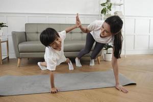 Young Asian Thai mother lovely trains her little son to exercise and practices yoga on living room floor together for healthy fitness and wellness, happy domestic home lifestyle on family weekends. photo