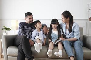 Asian Thai family, adult dad, mum, and children happiness home living relaxing activities and reading book together, leisure on sofa in white room house, lovely weekend, wellbeing domestic lifestyle. photo