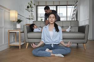 Young Asian Thai mother sits on living room floor, meditates, and practices yoga for health and wellness when husband and son tease together on sofa, happy domestic home lifestyle on family weekend. photo