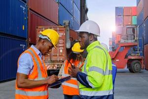 los trabajadores afroamericanos y los equipos con uniformes de seguridad y cascos usan walkie-talkies, trabajan en grúas logísticas con pilas de contenedores, control de carga, envío de mercancías e industria de transporte de carga. foto