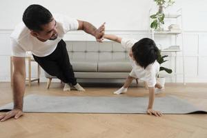 Young Asian Thai father lovely trains her little son to exercise and practices yoga on living room floor together for healthy fitness and wellness, happy domestic home lifestyle on family weekends. photo