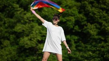 mujer joven de blanco con gafas de sol y nudos superiores sostiene la bandera del orgullo y la ondea en el viento frente a los árboles del parque ata video