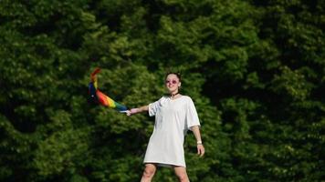 Young woman in white with sunglasses and top knots holds Pride flag and waves it in the wind in front of trees ata park video
