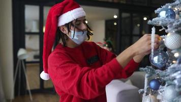 jeune homme en chemise rouge et bonnet de noel décore un arbre de noël tout en portant un masque video