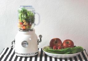 white vintage blender or smoothie maker  with a plate of vegetables, tomatoes and apples on  black and white stripe table cloth and white wall. Healthy drink making. photo