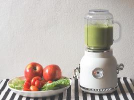 white vintage blender or smoothie maker  with a plate of vegetables, tomatoes and apples on  black and white stripe table cloth and white wall. Healthy drink making. photo