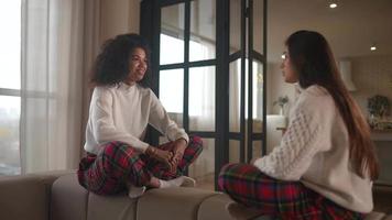 dos mujeres jóvenes se sientan en un sofá con suéteres a juego y pijamas a cuadros hablando entre sí video