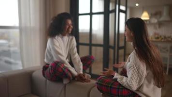 dos mujeres jóvenes se sientan en un sofá con suéteres a juego y pijamas a cuadros hablando entre sí video