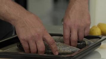 Male hands prepare several fish for baking on a baking sheet. Add oil, salt and spices to the fish. Cooking process.