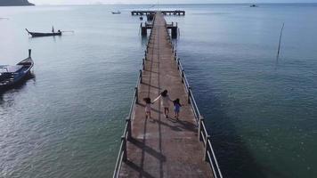 Aerial view from a drone of a happy Asian family of mother and daughters having fun running along the pier in a tropical sea during summer vacation. Travel and vacation concept. video