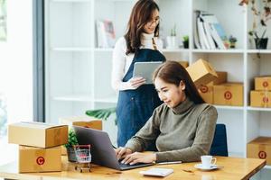 dos mujeres felices que venden productos en línea inician una pequeña empresa usando una computadora portátil y una tableta para calcular precios y prepararse para el franqueo. foto