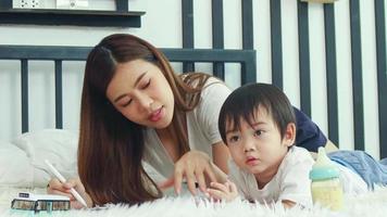 asiatico madre e figlio felicemente giocando nel letto insieme.madre insegna compiti a casa e insegna bambini per disegno. video
