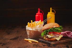 Homemade burger with beef, cheese and onion marmalade on a wooden board, fries in a metal basket and sauces. Fast food concept, american food photo