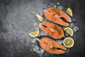 Fresh salmon steak with herbs and spices lemon rosemary on black plate background Raw salmon fish fillet and ice for cooked steak seafood photo