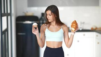 une jeune femme en tenue de sport tient un cupcake et un croissant et regarde d'avant en arrière les deux video