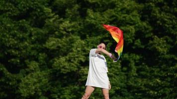 Young woman in white with sunglasses and top knots holds Pride flag and waves it in the wind in front of trees video