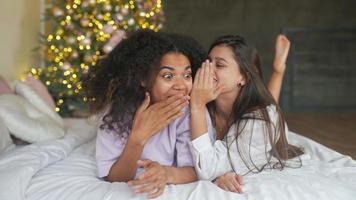 Two young women sit on bed in pajamas and talk with Christmas tree in background video