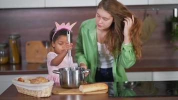 Young woman with long red curly hair laughs and looks at the camera while she and a younger girl cook together in a kitchen