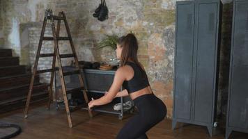 Athletic young woman does squats wearing black leggings and crop top before brick wall and lockers and ladder video