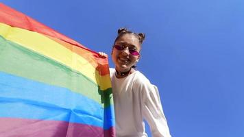 mujer joven con gafas de sol y nudos superiores sostiene la bandera del orgullo y da el signo de la paz en un ángulo de cámara bajo
