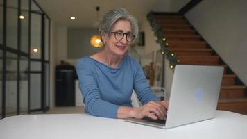 Woman with gray hair and glasses types on laptop before looking up to make eye contact as lights flicker in the background video