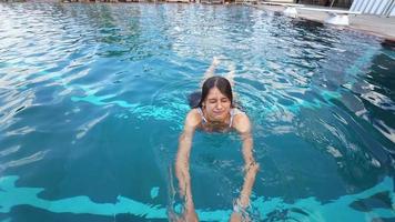 Young woman in a pool looks at and swims toward camera laughs and smiles while others lounge poolside video