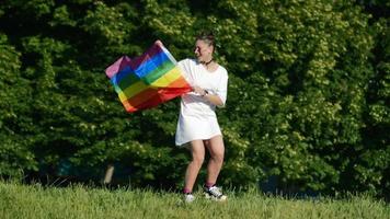 mujer joven de blanco con gafas de sol y nudos superiores sostiene la bandera del orgullo y la ondea en el viento frente a los árboles video