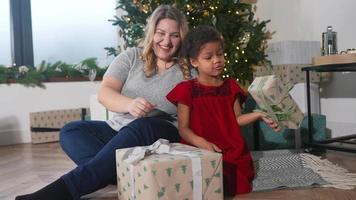 Young girl in red dress wonders about a wrapped gift in front of a lit decorated Christmas tree while adult woman family member laughs video