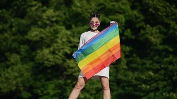 Young woman in white with sunglasses and top knots holds Pride flag and waves it in the wind in front of trees ata park video