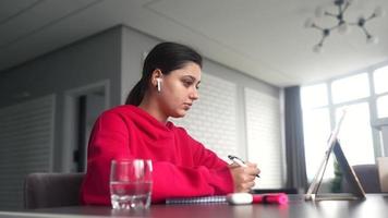jovem em um moletom rosa brilhante senta-se em uma mesa com fones de ouvido de um tablet enquanto escreve em um caderno com uma caneta