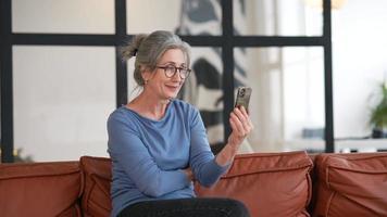 mujer con cabello gris y anteojos se sienta en un sofá usando un teléfono inteligente para chatear por video