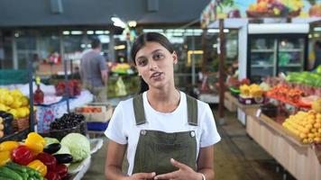 Young woman in green overalls talks to camera in the produce section of a grocery store video