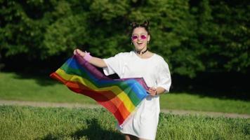 Young woman in white with sunglasses and top knots holds Pride flag and waves it in the wind in a park video