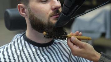 Barber uses brush and blow dryer to dry and shape man's beard after a shave