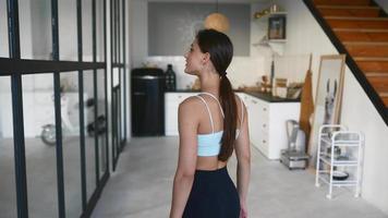 Fit young woman in leisure active wear looks at camera while showing us around an open sunny living space video
