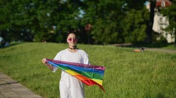 Young woman in white with sunglasses and top knots holds Pride flag and waves it in the wind in front of trees video